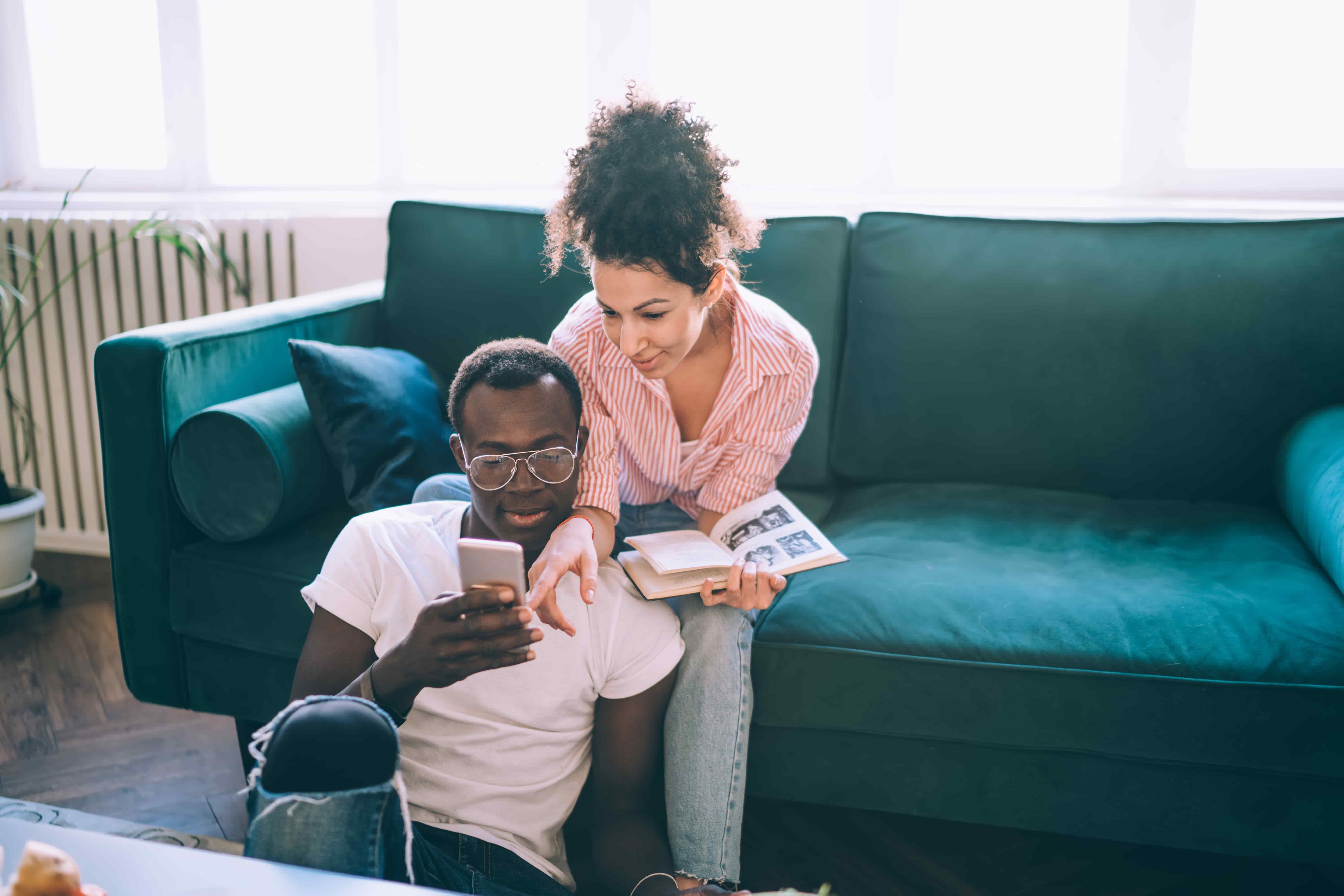 Enthusiastic curly woman with book pointing on phone for showing news to black concentrated husband while relaxing together in cozy house